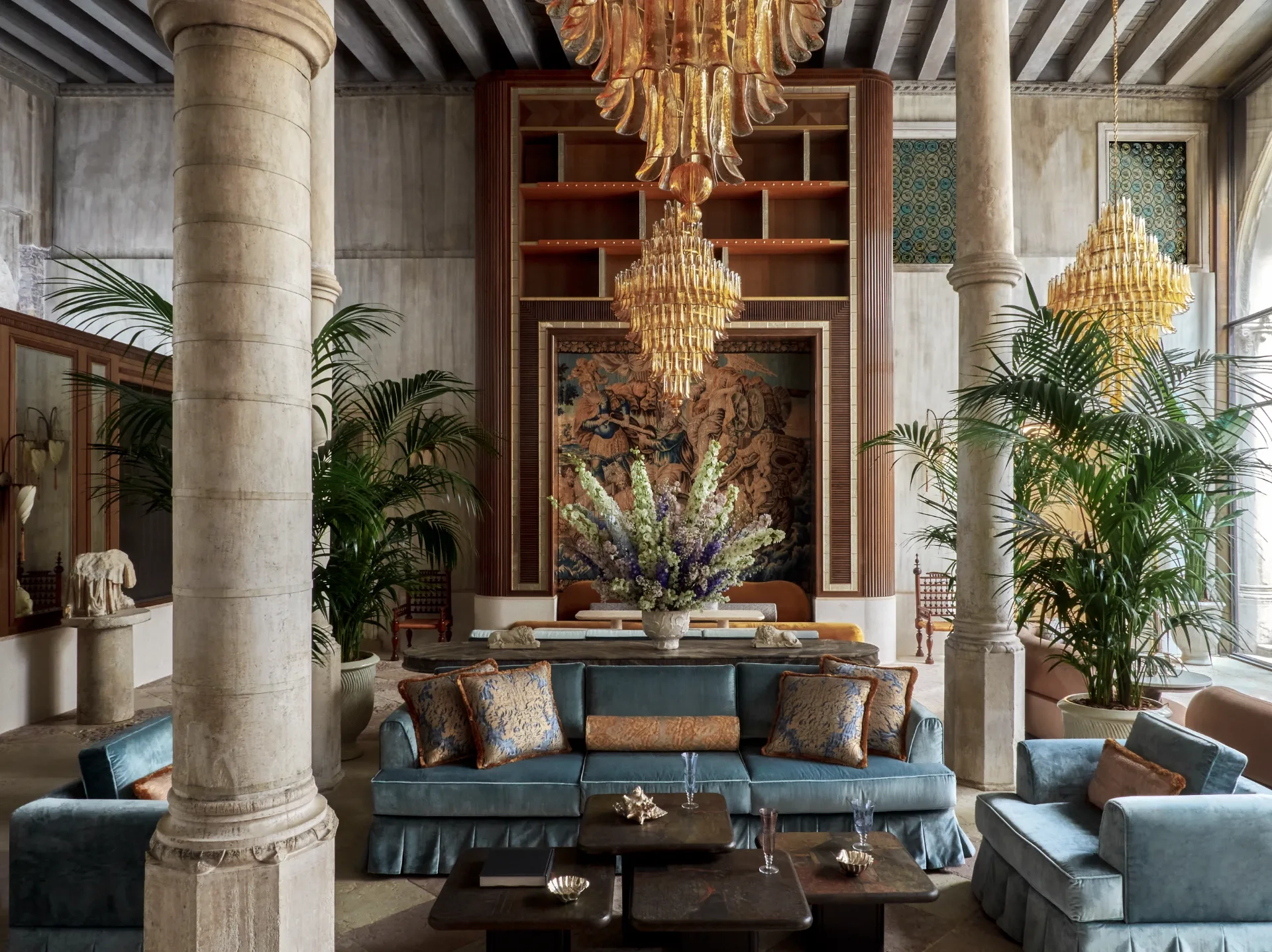 The lobby at Orient Express Venezia features stone columns, chandeliers, a blue couch and large floral arrangements
